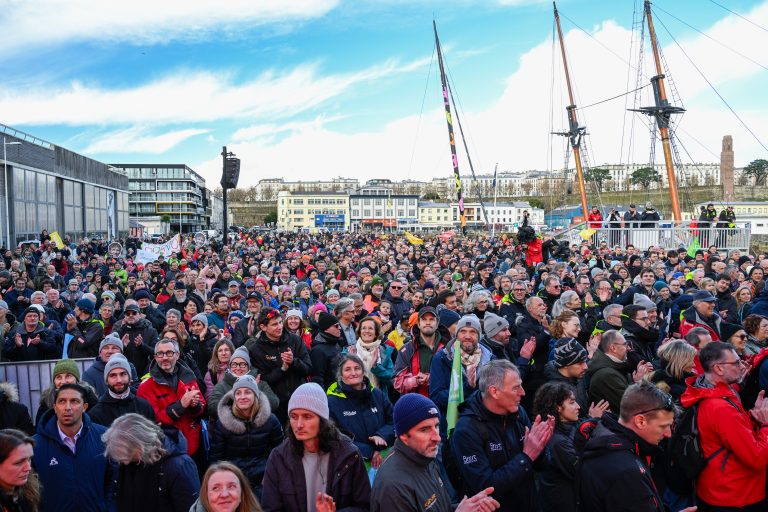 Foule nombreuse de spectateurs applaudissant sur un quai de port en hiver, m&acirc;ts de voiliers en arri&egrave;re-plan.